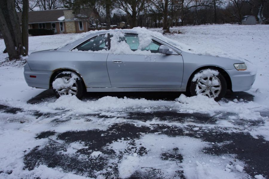 A snow-covered car sits in the middle of a driveway, waiting to be taken on a drive. With snow on the tires and on the roads, people need to give themselves extra time to slow to a stop.